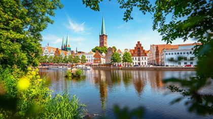 View of the historic city centre of Lübeck with colourful façades, tall church spires, and a river in the foreground, framed by green trees on a sunny day.