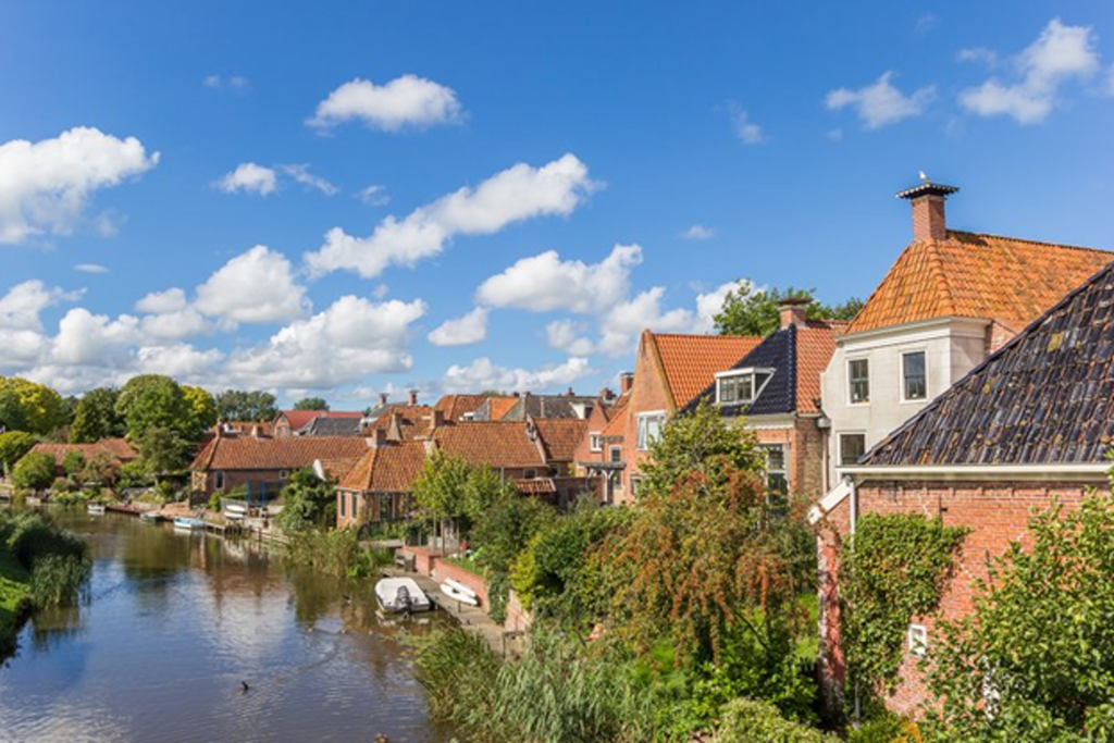 Charming canal-side village with traditional red-roofed houses along the Groningse Merenroute in the Netherlands, under a bright blue sky.
